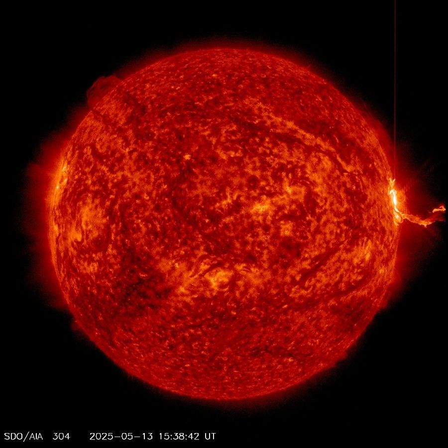 The Sun appears in shades of red, with darker and brighter areas scattered around. On the far right edge of the Sun is a very bright area, with a long filamentary structure extending outward from it, toward the right.