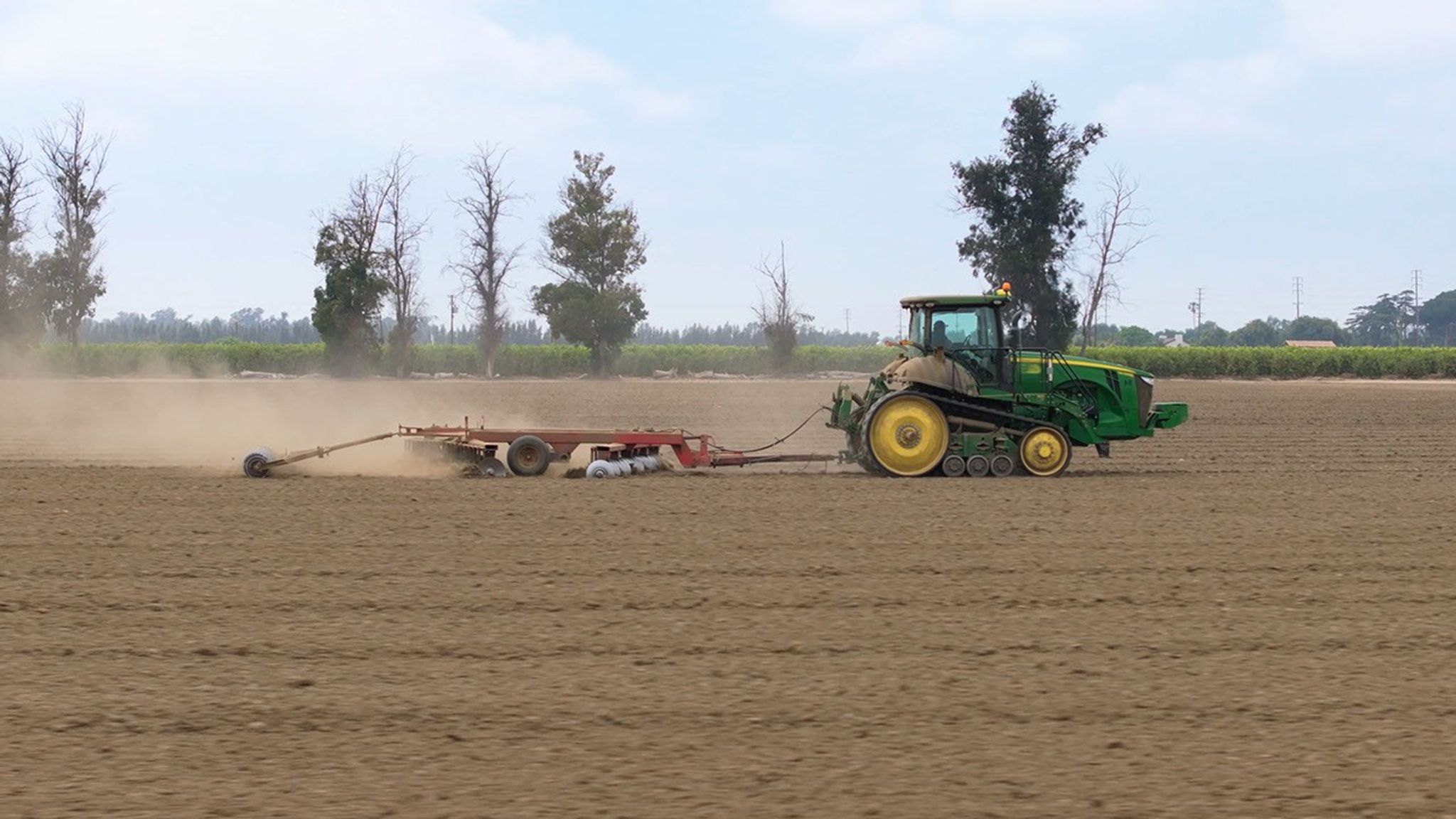 A green tractor with yellow wheels pulls a red trailer across a flat, dirt-covered field with some trees, a field of green plants, and a partly cloudy sky in the background.