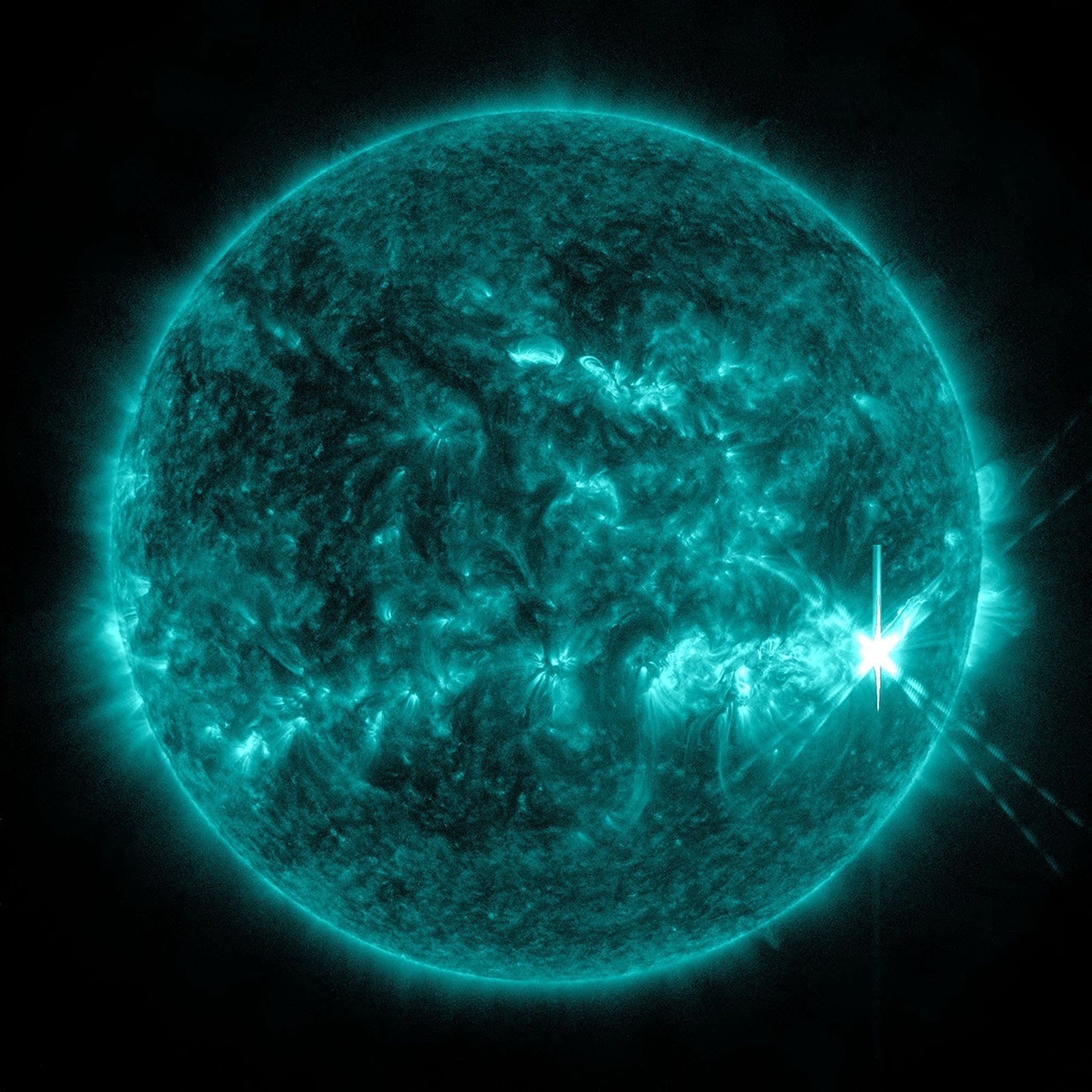 A bright turquoise image of the sun, displaying swirling surface patterns and a large solar flare erupting off the right edge, captured in ultraviolet light against a black background.