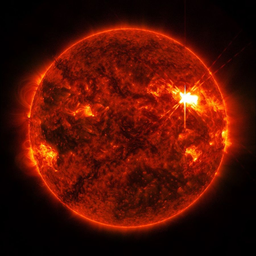 An extreme ultraviolet view of the Sun shows the bright flash of a solar flare on the Sun’s upper right side. The rest of the surface is mottled with darker and lighter regions, and faint loops of solar material can be seen extending off the Sun’s edges.