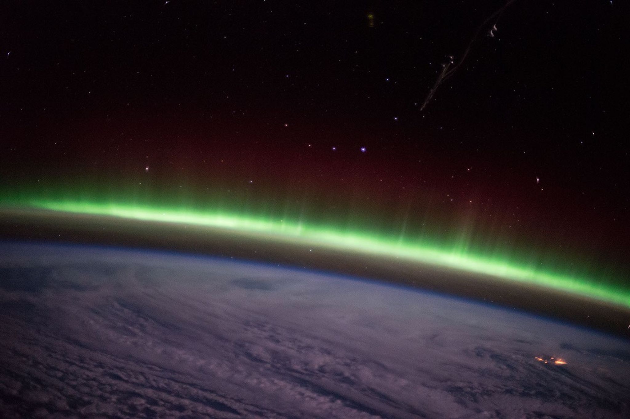 An arc of green aurora bends over the limb of Earth, which is covered in swirling clouds. A faint red haze appears above the green auroral arc against the black background of space and a couple dozen stars.