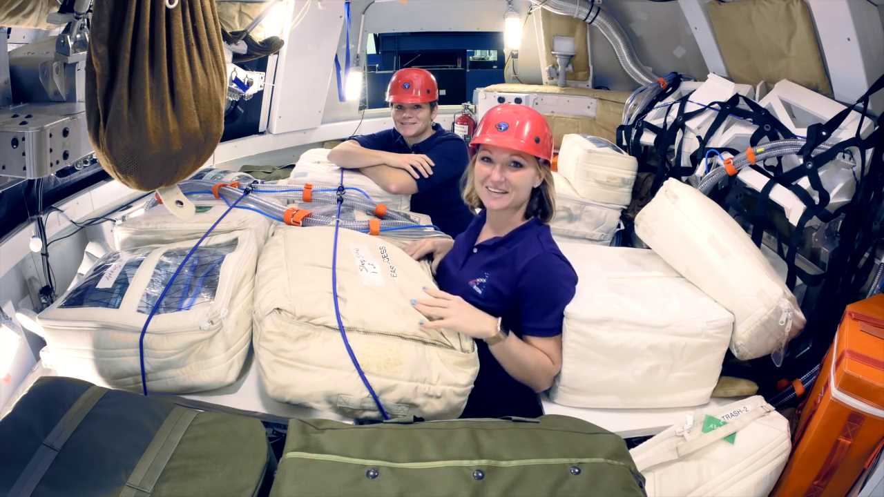 Two people wearing pink hard hats appear at center inside a large, round capsule, looking at the camera and smiling. They are surrounded by large, filled bags, some white and some green, and they appear as if they are partially buried by the bags, with only the upper half of their bodies appearing above the bags.