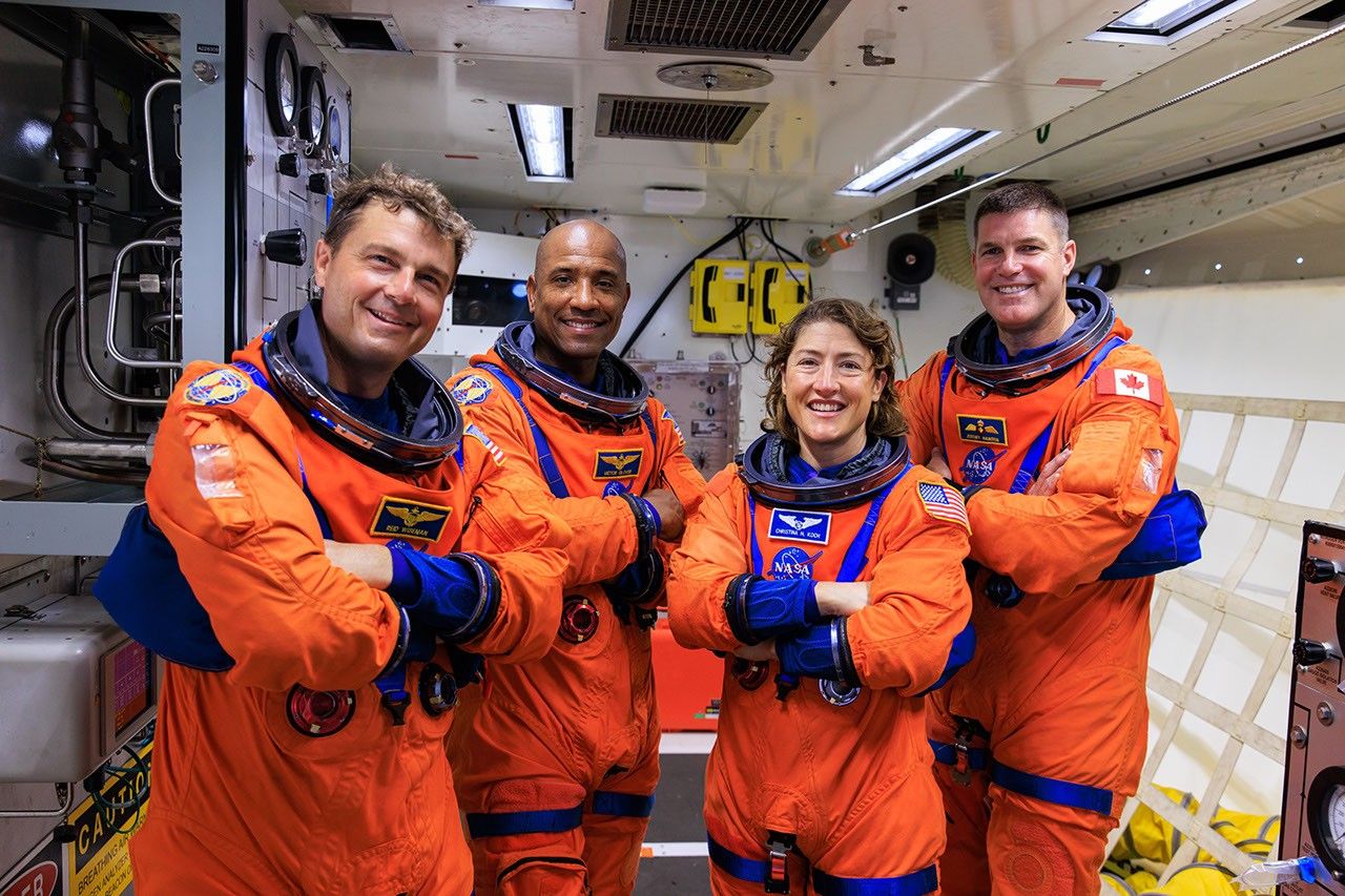 Four astronauts in bright orange flight suits stand smiling in the white room at Launch Pad 39B. The Artemis II crew members - NASA astronauts Reid Wiseman, Victor Glover, Christina Koch, and CSA astronaut Jeremy Hansen - are lined up side-by-side during a ground systems test at Kennedy Space Center.