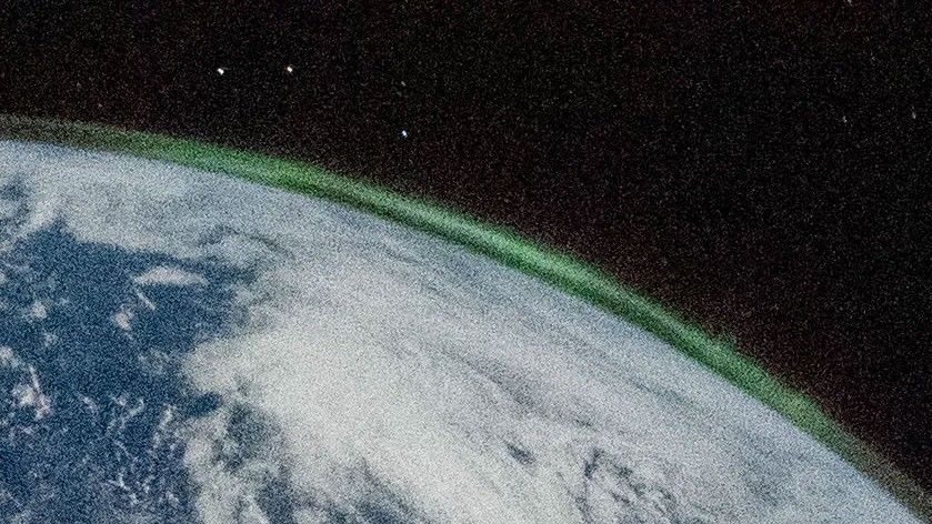 A small segment of Earth’s horizon. Earth fills the lower left corner. Green aurora light up the outer edge of the atmosphere. The aurora-capped atmosphere looks like a very thin shell.