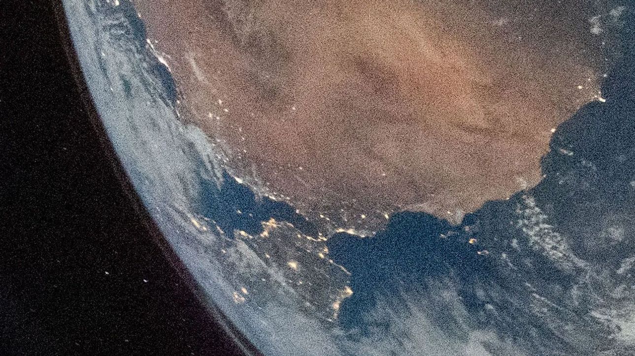 A segment of Earth's horizon, with what looks like a land bridge connecting two continents. Its edges are dotted with lights. Earth fills the upper right corner of the image. In the middle of the peninsula closest to the horizon, an arrow points to a bright spot.