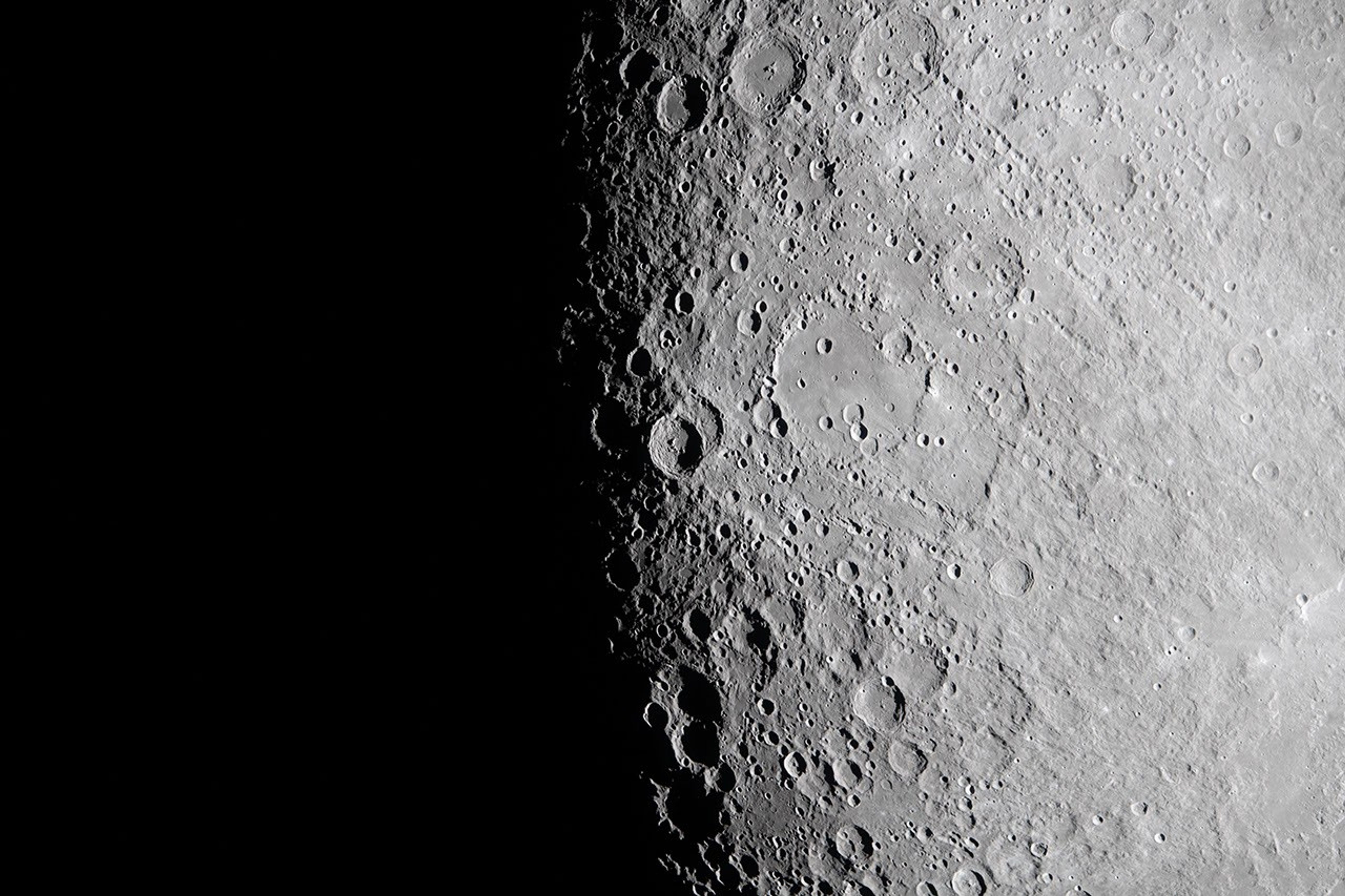 View of the Moon’s surface at the lunar terminator, where bright sunlight meets deep shadow. The right side of the image shows a highly detailed, grey, crater‑covered landscape, with craters of many sizes creating a textured appearance. Toward the left, the surface transitions sharply into complete darkness, marking the boundary between the illuminated and unlit regions of the Moon.