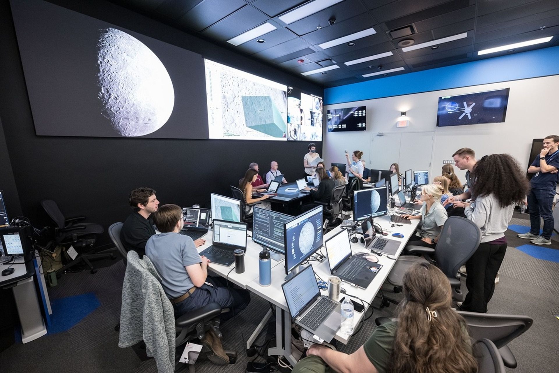 Group of people seated at a table with many computers and monitors. There are large screens on the wall of the Moon.