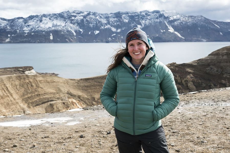 Kelsey Young smiles while standing outside in rugged terrain. Kelsey is wearing a knit cap and puffer jacket with the wind blowing her hair. She is standing on rocky soil with patches of ice. Behind her is a ridge of tan rock and a calm lake. Across the lake, rocky mountains with patches of snow and ice rise into the cloudy gray sky.