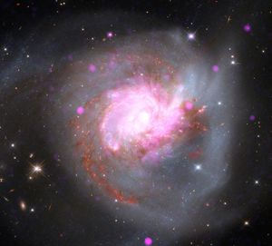 A pair of colliding, gas-rich spiral galaxies merging into a single massive system. Chandra’s X-ray data (pink) isolates point sources and shock-heated gas, Hubble’s optical light (blue and white) captures tidal tails of stars, and Webb’s infrared vision (red and orange) cuts through the dust to show hidden star formation. Galaxy collisions like this offer a preview of our far future, showing what might happen if our own Milky Way collides and merges with the nearby Andromeda Galaxy.