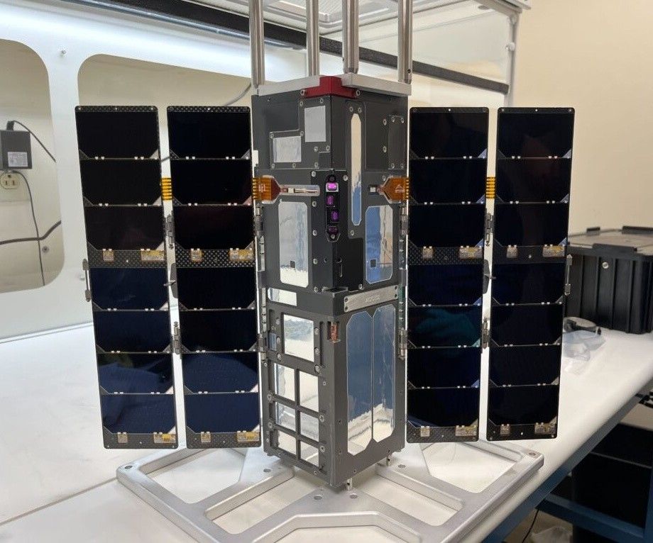 A small satellite with solar panels extended on either side sits atop a lab bench.