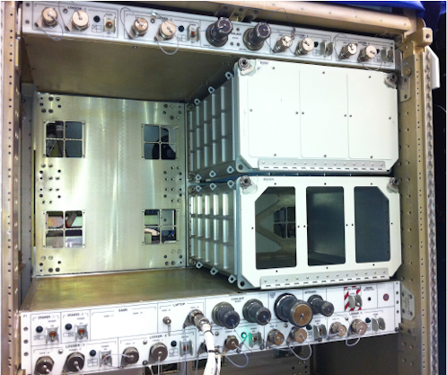A close-up view of an equipment rack, likely in a spacecraft or laboratory, with several modules installed. On the right, two white, box-like modules are stacked vertically, with the lower one showing an open front revealing internal compartments. The left side of the rack is an empty bay with a metallic back panel featuring several square openings. The top and bottom sections of the rack are populated with numerous knobs, connectors, and switches, and labels such as "LOCKER," "SPEAKER," "RAMS," and "LAPTOP" are visible.