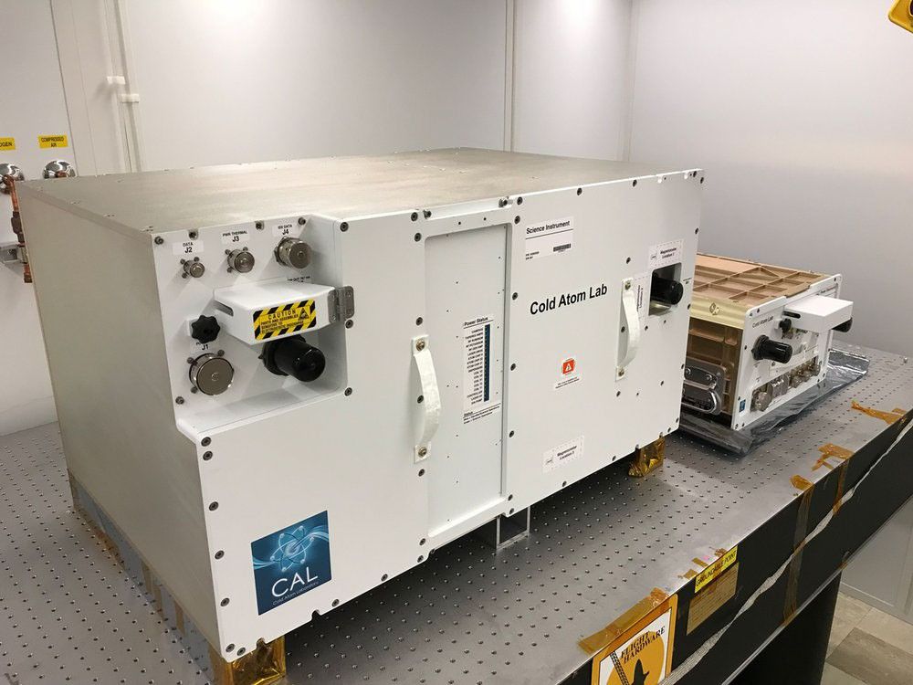 A large, white, rectangular scientific instrument labeled "Cold Atom Lab" and "Science Instrument" sits on a dark, gridded laboratory table. The instrument has various connectors, knobs, and handles on its front surface. To the right and slightly behind it, a smaller, metallic-colored piece of equipment also rests on the table. The setting appears to be a cleanroom or laboratory with plain white walls.