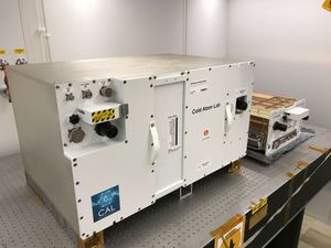 A large, white, rectangular scientific instrument labeled "Cold Atom Lab" and "Science Instrument" sits on a dark, gridded laboratory table. The instrument has various connectors, knobs, and handles on its front surface. To the right and slightly behind it, a smaller, metallic-colored piece of equipment also rests on the table. The setting appears to be a cleanroom or laboratory with plain white walls.