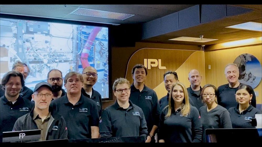 A group photo of the JPL Cold Atom Lab operations team, consisting of about fourteen men and women, posing together in what appears to be a control room. Many are wearing matching black polo shirts with the "Cold Atom Lab" logo. Behind them, a large screen displays an image of complex scientific hardware, and the JPL logo is prominently featured on the wood-paneled wall. The team members are smiling and looking at the camera.