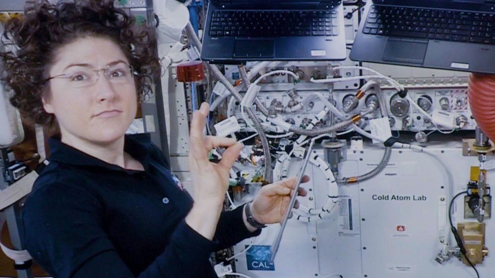 NASA astronaut Christina Koch, a woman with short curly brown hair and glasses, floats inside a module of the International Space Station. She looks toward the camera with a slight smile, making an "OK" hand gesture while holding a long, thin metal instrument. Behind her is a wall of complex scientific equipment, including the Cold Atom Lab, with numerous wires and two laptops mounted upside-down.