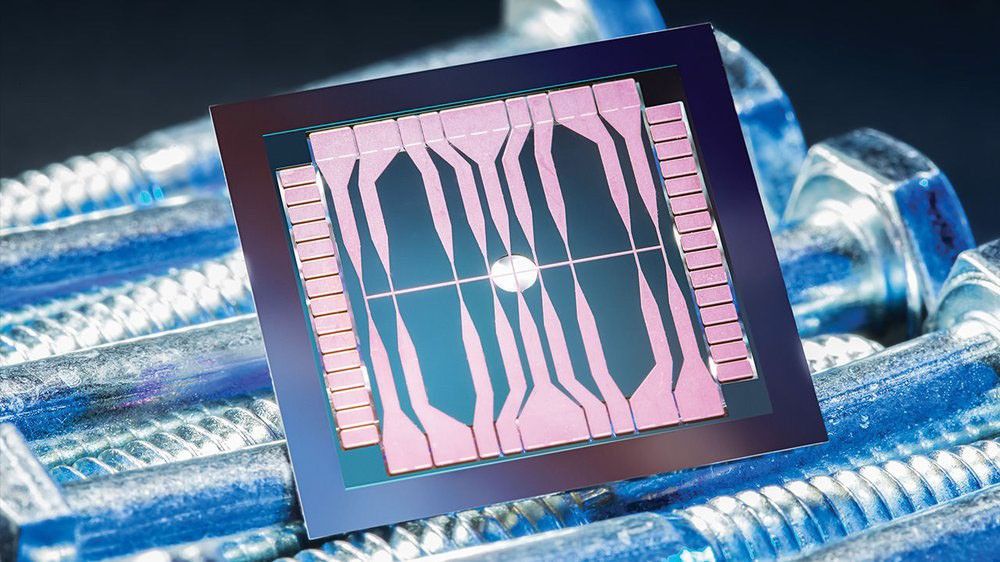 A close-up, high-resolution photograph of a square microchip with a dark frame, resting diagonally on a bed of several metallic bolts. The chip's surface features an intricate pattern of light pink or copper-colored traces and pads against a dark blue-green substrate. A prominent white circular feature is located at the center of the chip, with radiating lines. The bolts beneath are metallic and appear slightly textured.