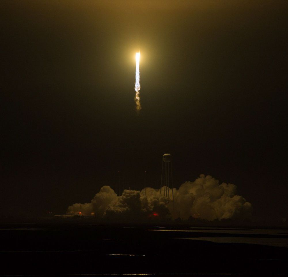 A rocket is captured mid-launch at night, ascending into a dark sky with a brilliant, fiery exhaust plume trailing behind it. The plume glows intensely yellow-white at its base and fades into a smoke trail. Below the rocket, the launchpad is obscured by a large cloud of white smoke and steam, illuminated by the launch. A tall, white water tower is visible on the right, silhouetted against the bright cloud. The foreground shows a dark, reflective surface, possibly water or a wet launch area.