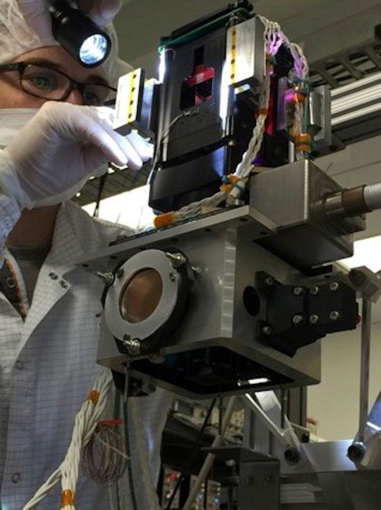 A scientist in a cleanroom suit, wearing a lab coat, hairnet, and gloves, leans over to inspect a complex piece of scientific equipment. They are holding a flashlight to illuminate the intricate, metallic instrument, which is covered in wires and features a prominent circular lens.