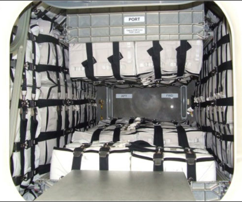 An interior view of a cargo bay, likely in an aircraft or spacecraft, filled with numerous rectangular white bags secured with black straps. The bags are stacked and strapped against the walls and floor of the compartment. Labels "PORT," "AFT," and "FWD" are visible on the interior walls, indicating directions within the cargo hold. The overall impression is one of tightly packed and organized storage.