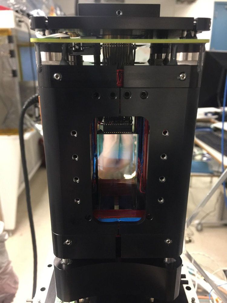 A close-up, eye-level photograph of a black, rectangular scientific instrument. A large rectangular opening in the front reveals internal components, including what appears to be a detector or sensor array. Visible inside are circuit boards and wires, with some reflections of light on the internal surfaces. The top and bottom sections of the instrument show metallic components and screws. The background is a blurred view of a laboratory setting with other equipment.