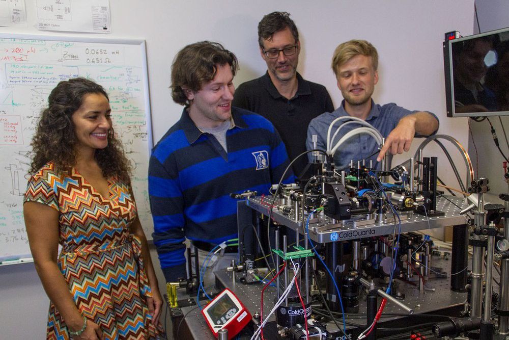 A group of four researchers, one woman and three men, gather around a complex piece of scientific equipment in a laboratory. The man on the right, with blond hair, points to a component of the intricate optical setup. The woman on the left, with long curly hair, smiles as she looks on. A whiteboard covered in handwritten equations and diagrams is visible in the background.