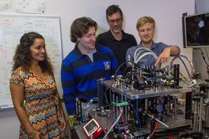 A group of four researchers, one woman and three men, gather around a complex piece of scientific equipment in a laboratory. The man on the right, with blond hair, points to a component of the intricate optical setup. The woman on the left, with long curly hair, smiles as she looks on. A whiteboard covered in handwritten equations and diagrams is visible in the background.