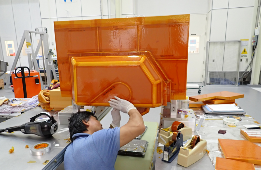 A man with black hair and a light blue polo shirt, his back to the camera, wearing protective gloves as he inspects an orange multi-layered box suspended overhead