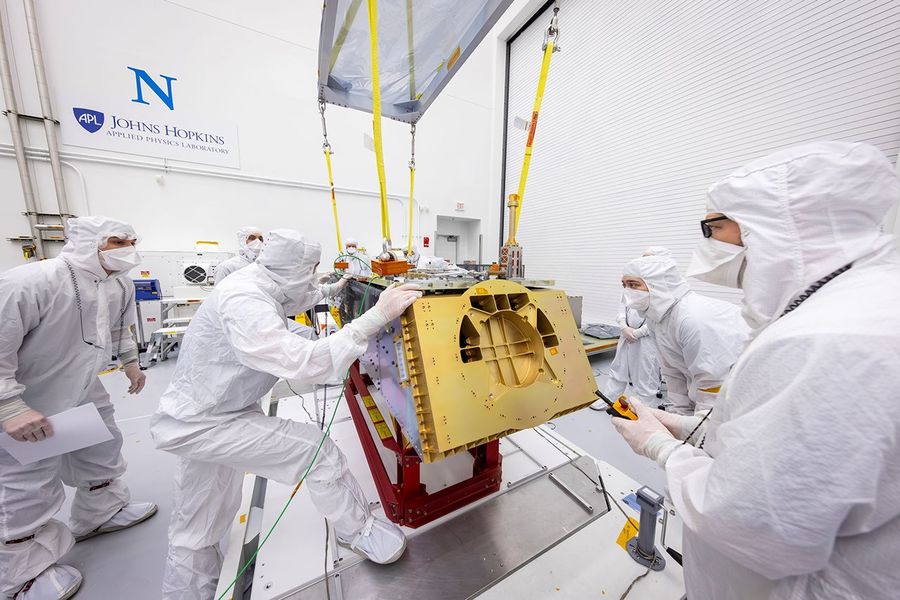 Technicians in cleanroom suits assemble panels of a NASA spacecraft in a brightly lit lab.