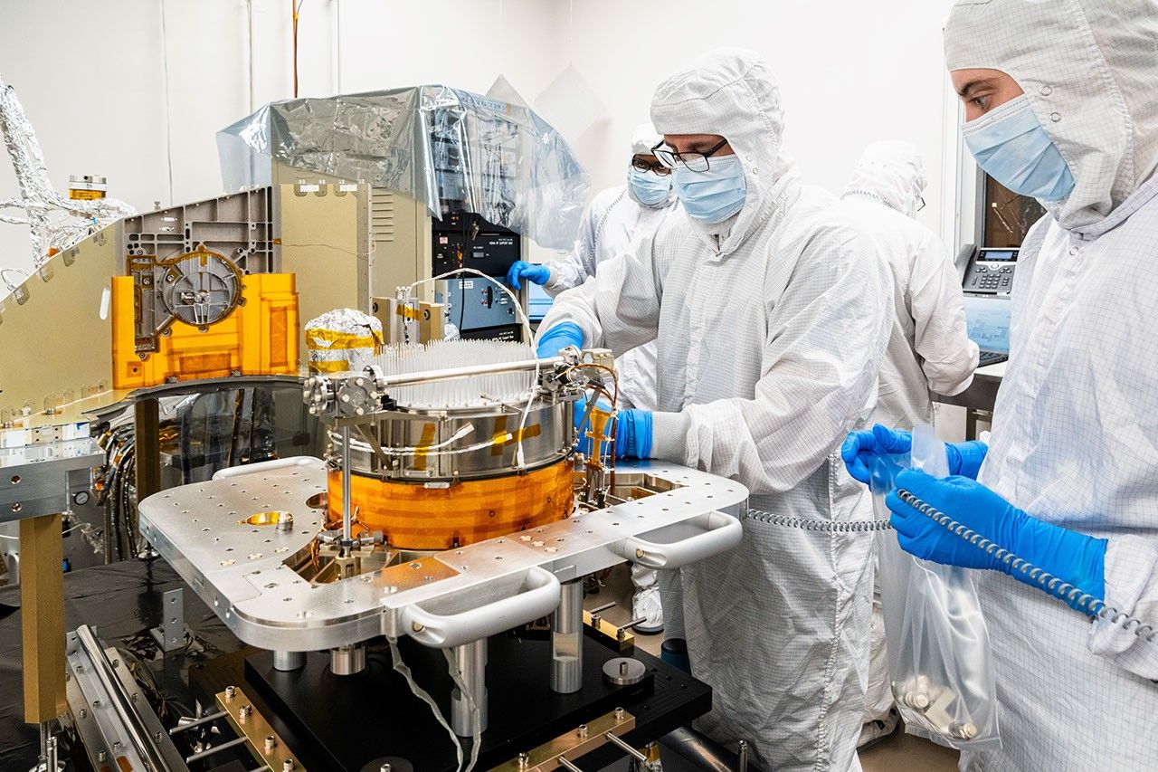 Cleanroom technicians assemble a complex spacecraft component, working around metallic hardware and wiring in a bright, sterile lab.