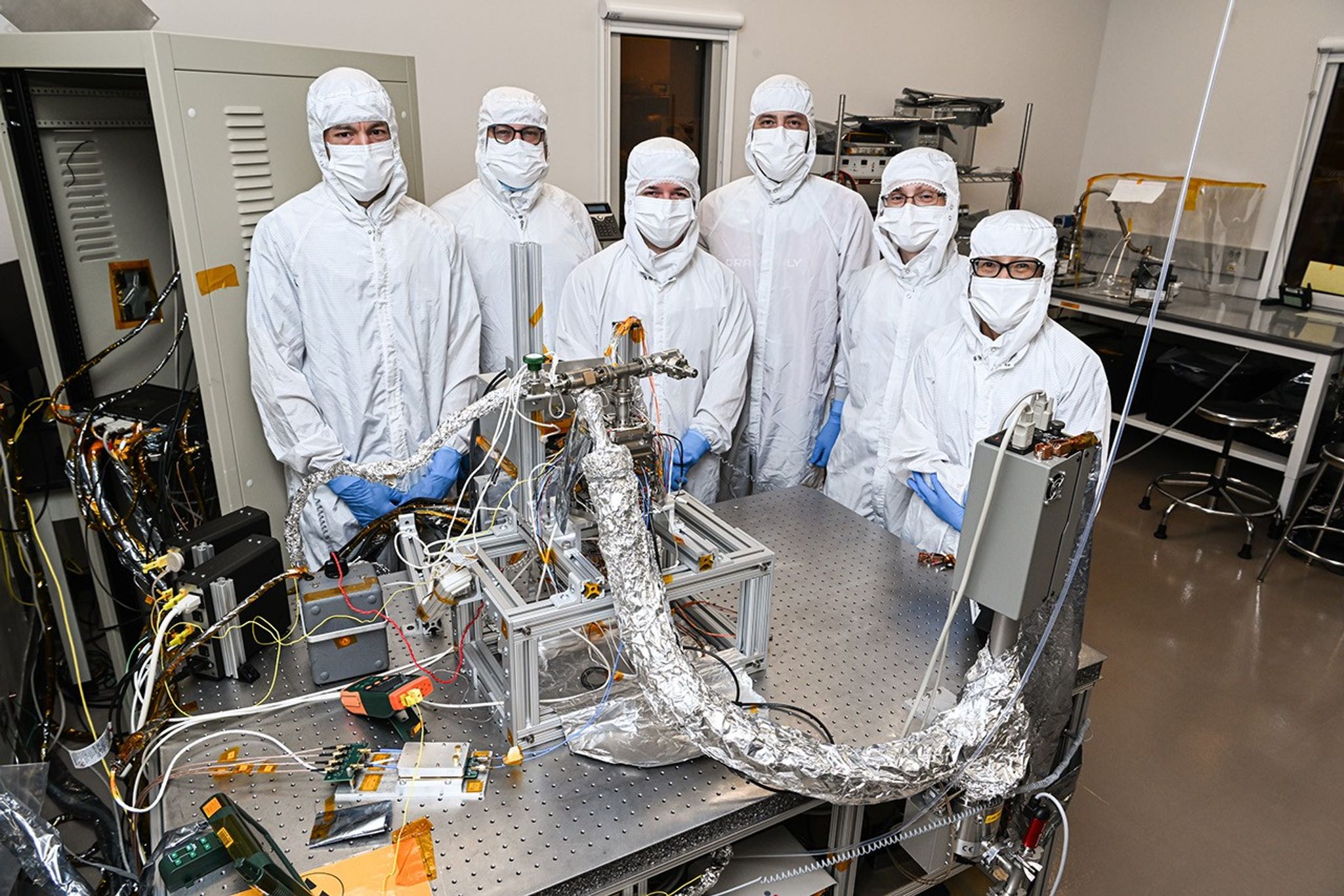 Six people in white masks and head-to-toe covered "bunny suits" pose behind a large tubular, foil-covered device leading into a see-through box with instruments on a metal table in a lab.
