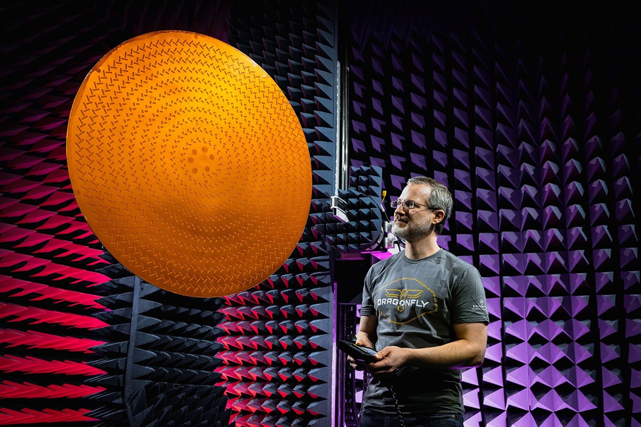 A spacecraft technician inspects a circular antenna in a foam-walled, soundproof test chamber.