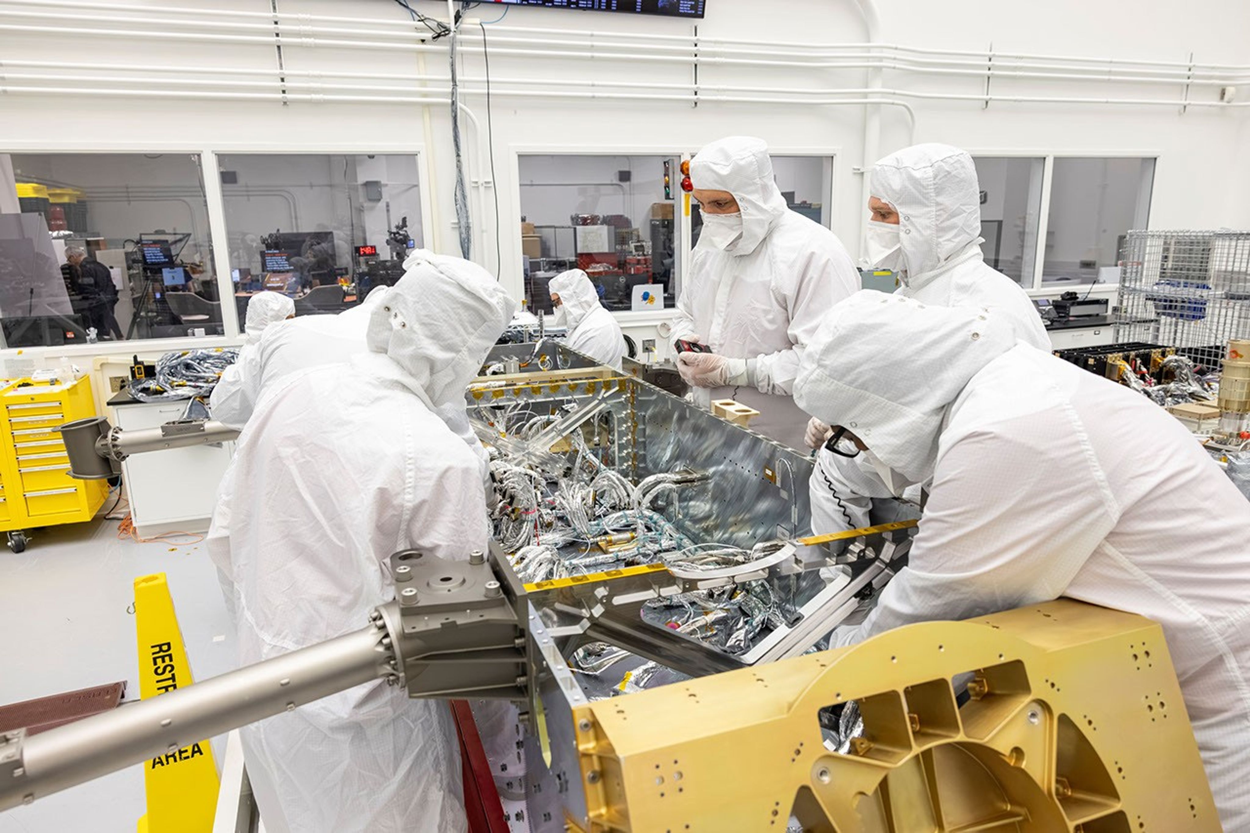 Workers attach electrical wiring to various spacecraft system components inside a clean room.