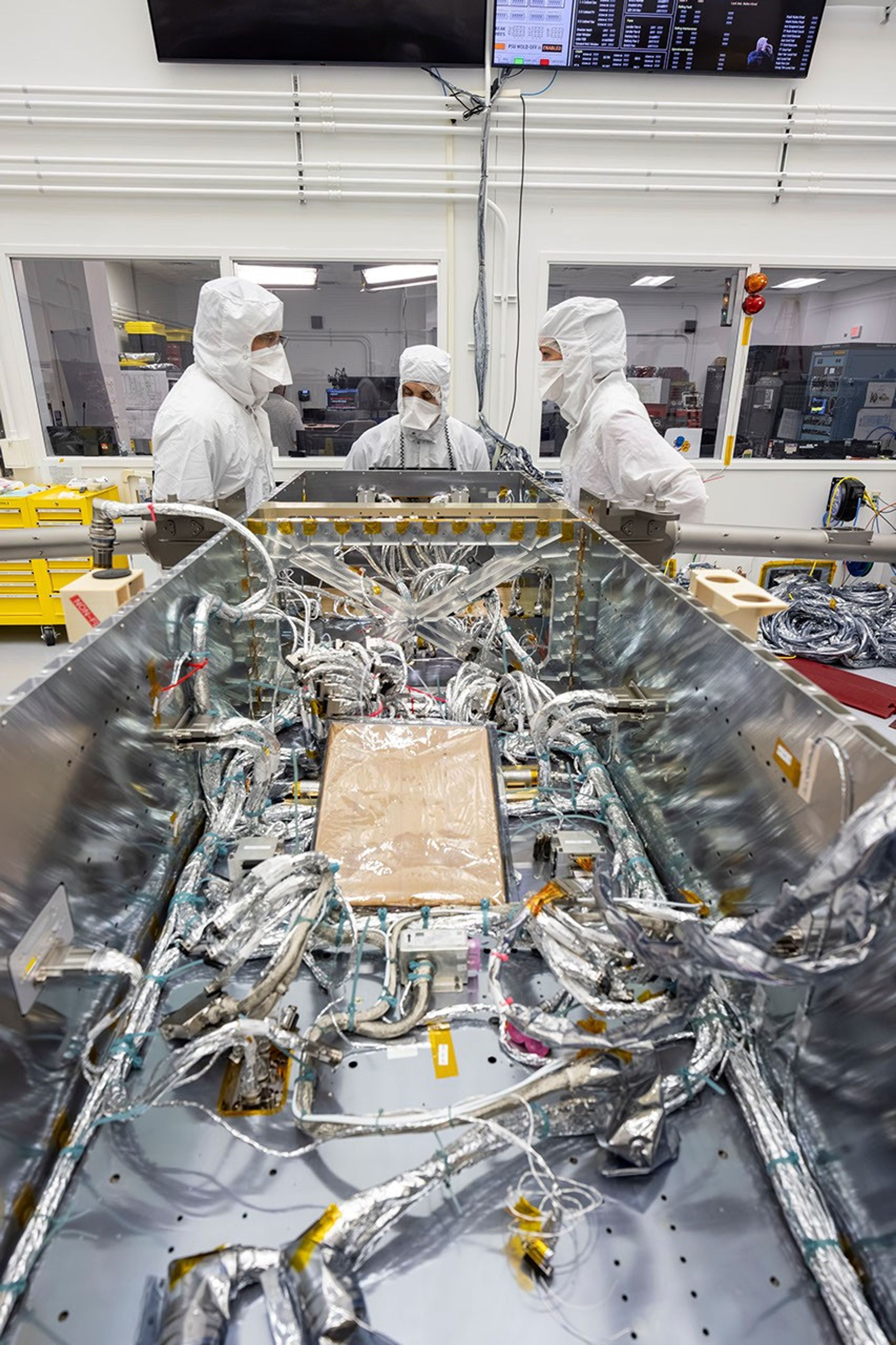 Technicians inside a clean room prepare to install electrical wiring to components inside of a spacecraft.