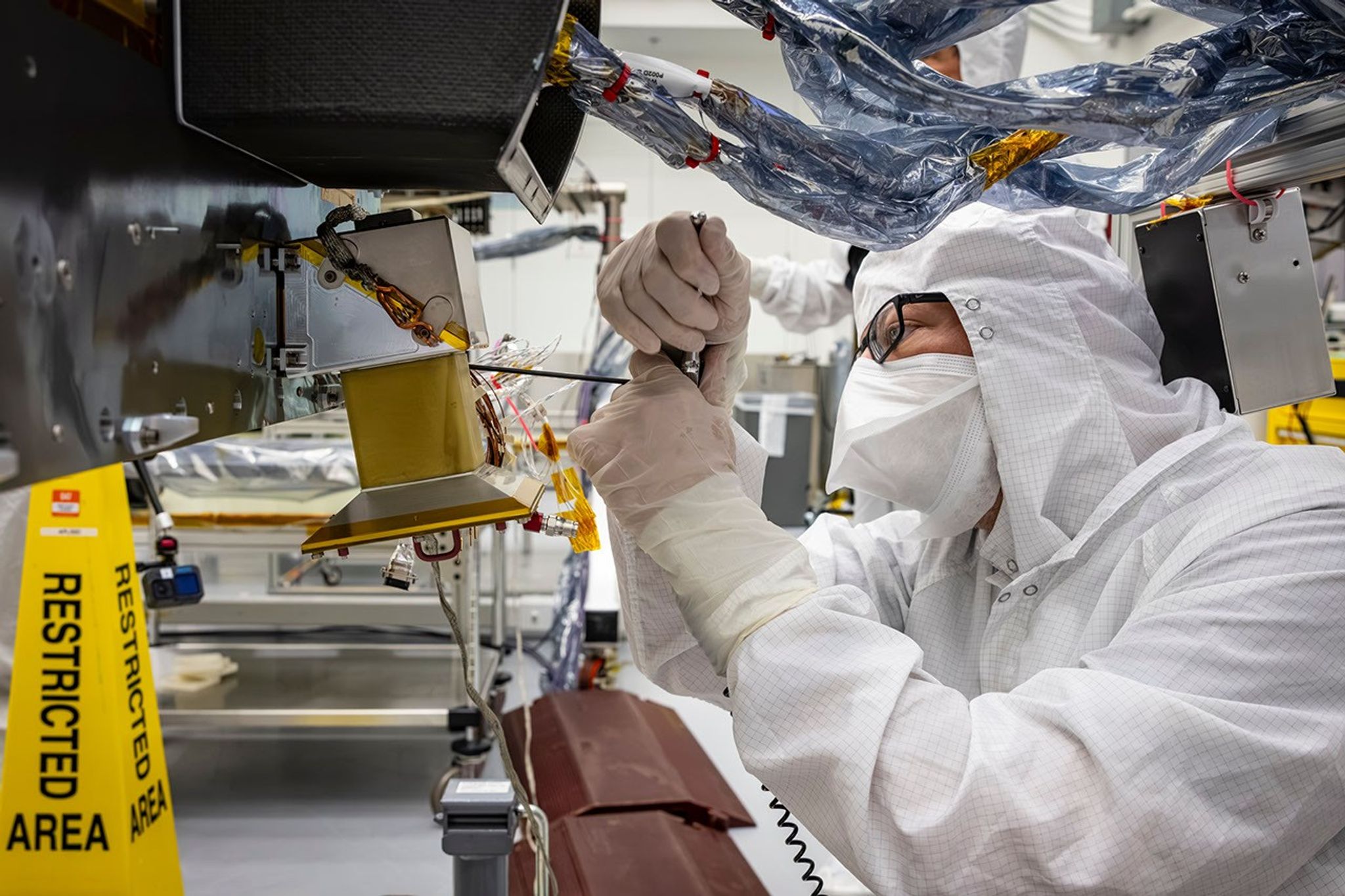 A technician in protective cleanroom gear carefully works on a spacecraft component surrounded by wiring and thermal insulation.