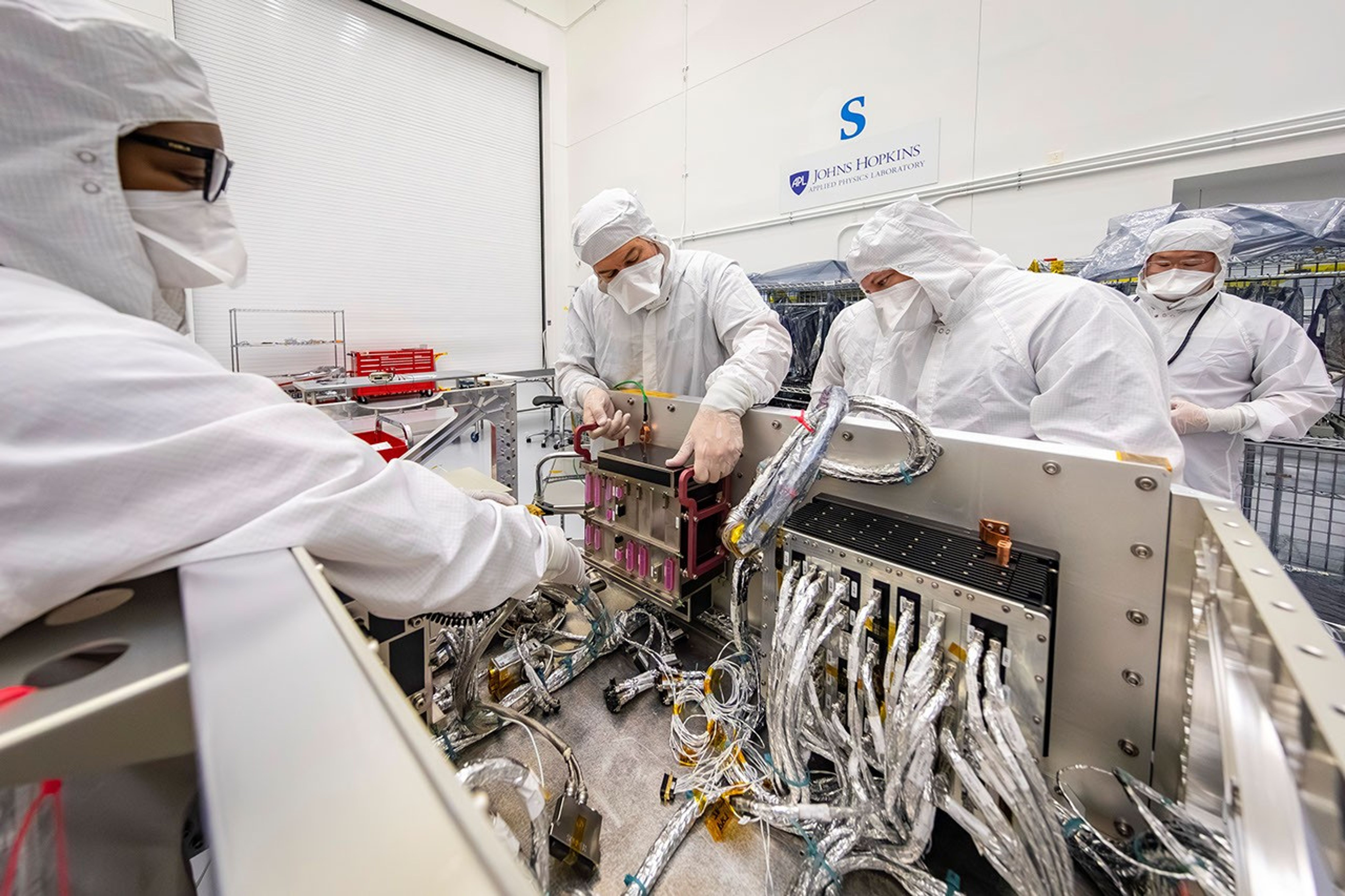At Johns Hopkins APL engineers in cleanroom suits connect electronics and computer boxes to a NASA spacecraft wiring system