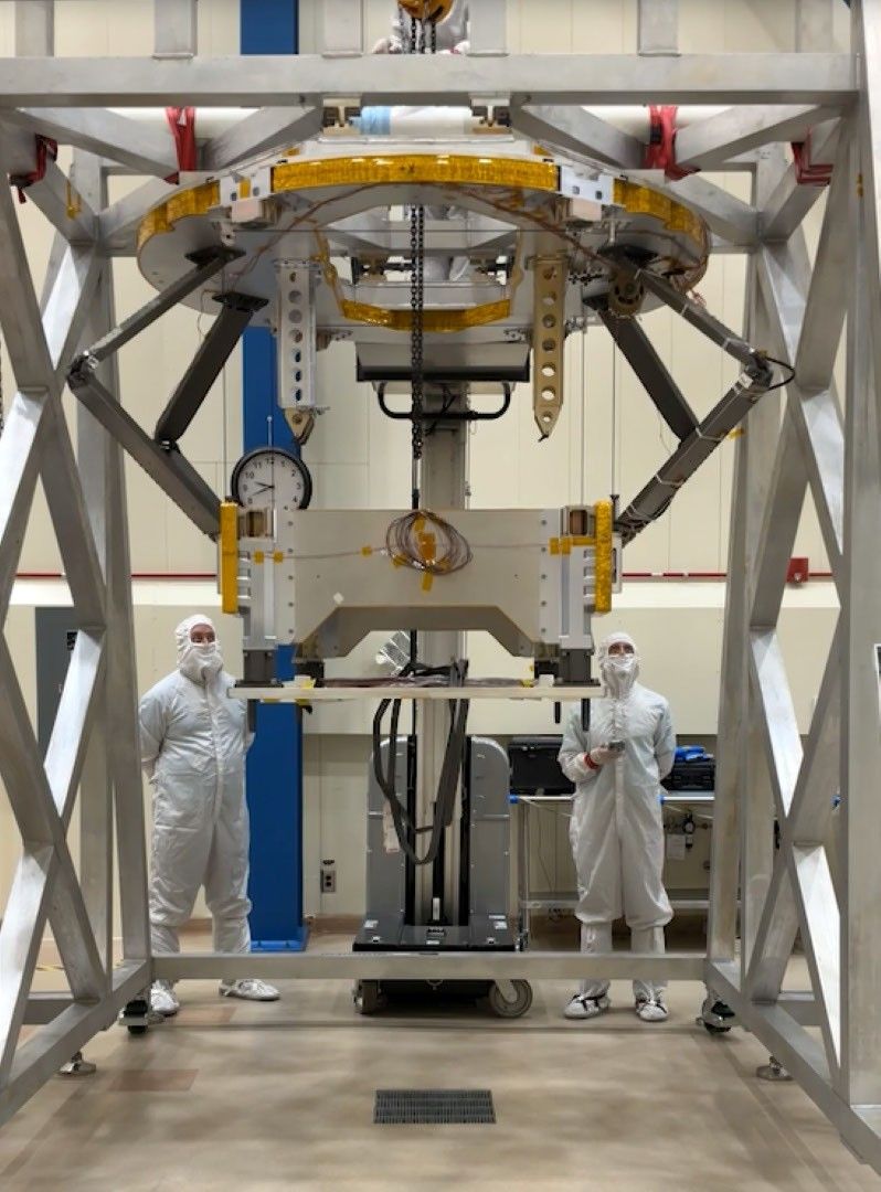 Two technicians in white cleanroom suits stand beside a large spacecraft testing apparatus supported by a metal frame in a laboratory.