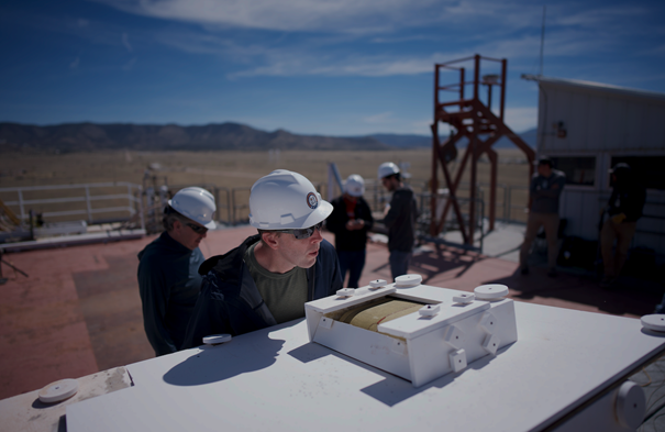 Workers inspect a large, box-shaped white instrument on a rooftop platform in the desert with mountains in the distance.