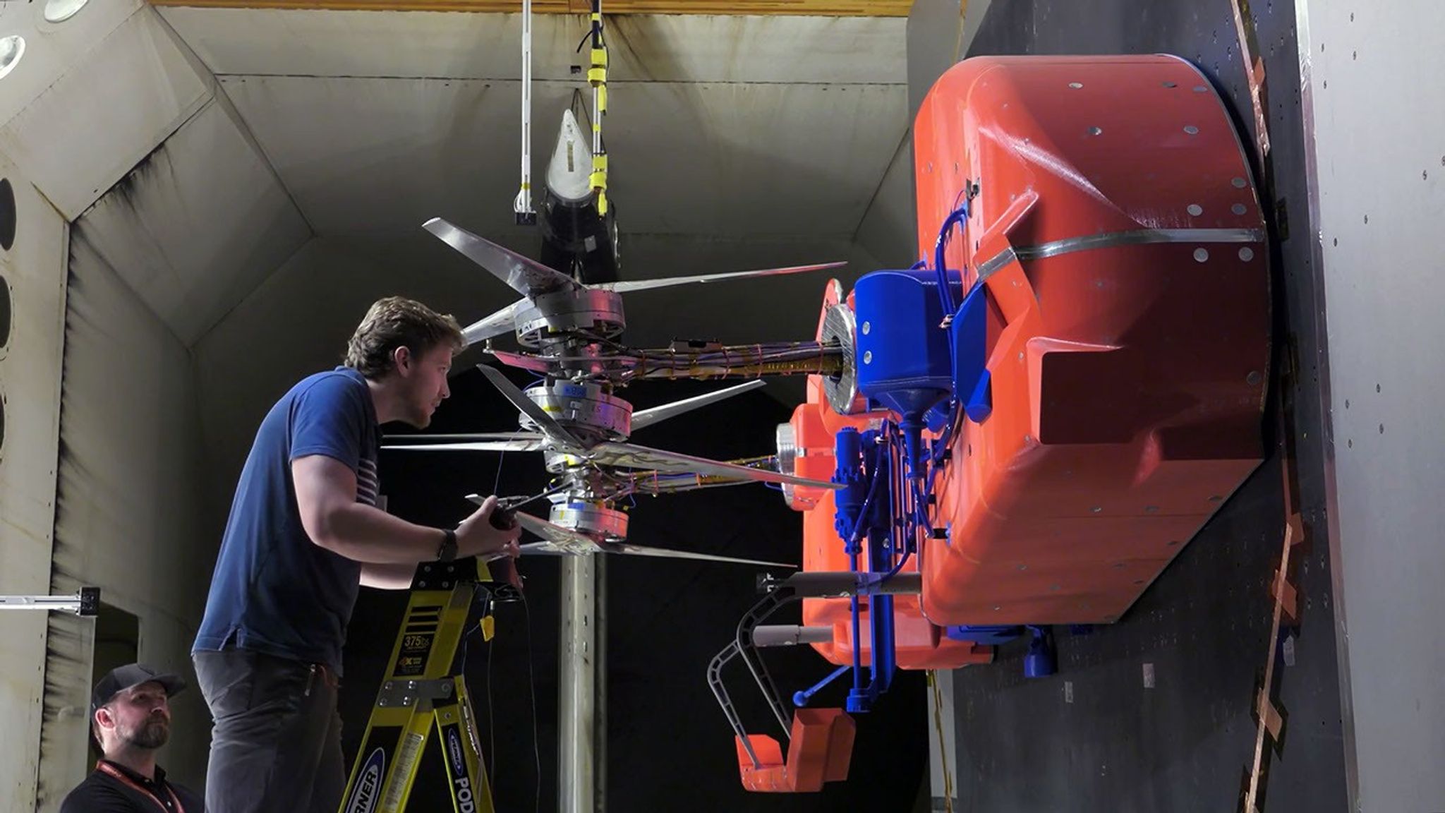 A man in a blue T-shirt on a ladder adjusting the rotor blades for the spacecraft model in the wind tunnel
