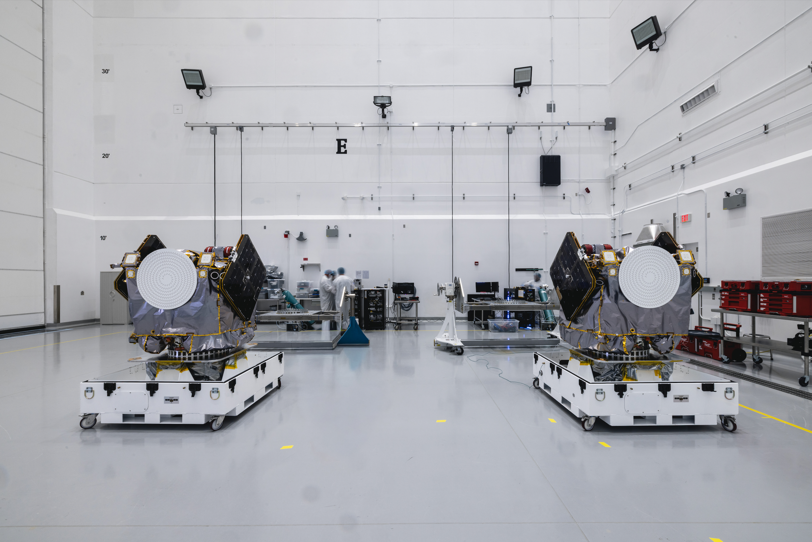 Two large spacecraft appear side by side inside a large, white-walled clean room..