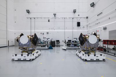 Two large spacecraft sit side by side, each on their own white, rectangular platform, inside a large white-walled clean room. Two people appear in the background.