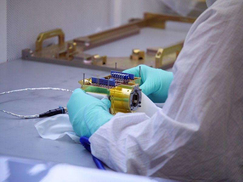 A small electronics device, with electronics visible at the top and gold metal around the bottom, is visible on a table. A person wearing white coveralls and light blue gloves is seen holding the device with both hands. Addition equipment is visible on the table surrounding the device, including a silver wire.