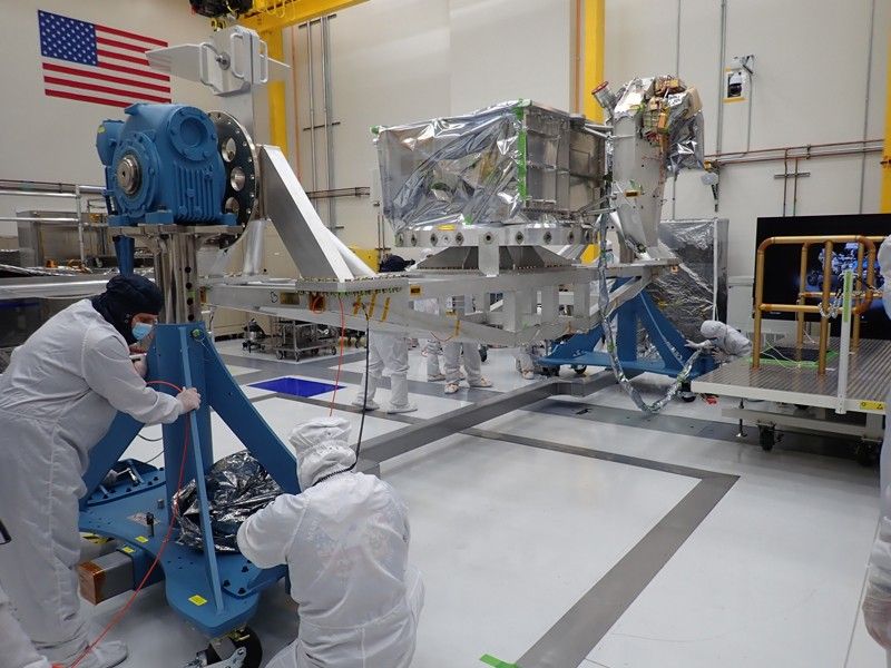 Two major pieces of flight hardware for the Europa Clipper spacecraft are attached to a flotron machine in a cleanroom. The hardware is visible in the rear middle of the images. The flotron is a large structure that allows the hardware to be rotated and moved for assembly purposes. The flight hardware consists of a large aluminum vault, which is shaped like a box. Within the vault, sensitive electronics will live, protecting them from radiation in Jupiter's environment. Attached the vault is a metal deck, called the nadir deck. Attached to this metal deck are multiple instrument sensors and cameras, which will allow the spacecraft to conduct science. Four engineers in full body coveralls and masks are seen working on flotron. An American flag is visible in the top left of the image.