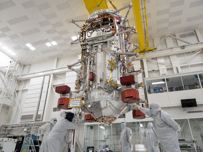 In the center of the image, the main body of the Europa Clipper spacecraft hangs from a yellow crane in a large, multistoried cleanroom with white walls and a white ceiling. Six engineers in full-body coveralls and masks stand around the base of the main body as it hangs from the crane, with some working on the main body. In the distance, a viewing gallery is visible with four people watching the activities taking place.