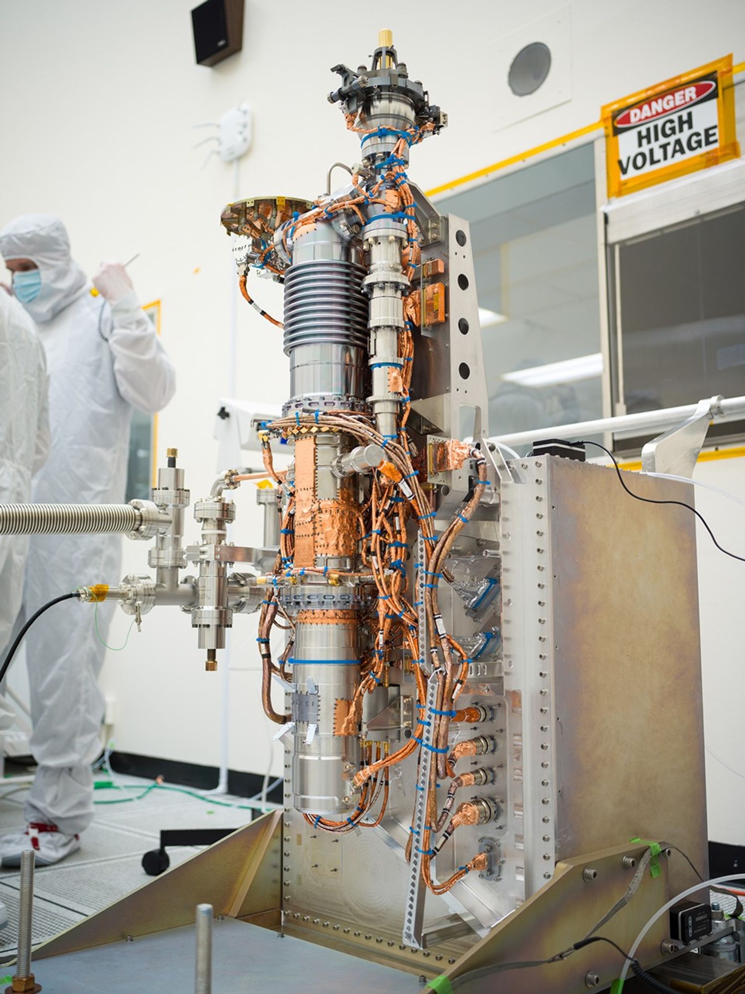 Europa Clipper’s mass spectrometer takes up the middle of this image. The instrument consists of a long cylinder, several feet tall and a couple of inches in diameter. It takes the shape of an automobile spark plug, with a series of ridges visible at the top. Copper plating and wires extend from the instrument in multiple areas. The instrument is installed on a supportive structure. Engineers are visible in the background working.