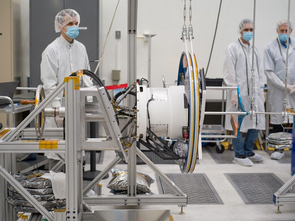 Three workers in white lab coats, cleanroom bouffant caps, and blue face masks watch as Europa Clipper’s magnetometer boom is tested. Part of the equipment holding the boom can be seen in front of the workers.