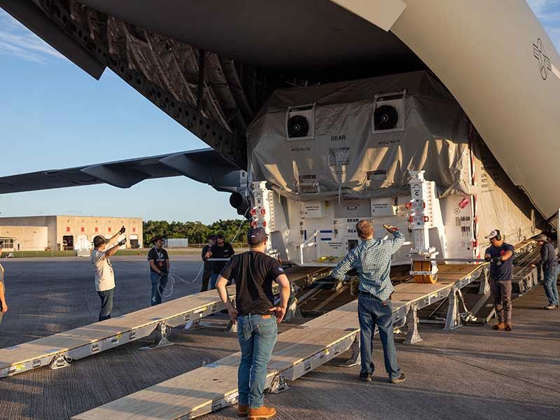 The back of a green transport aircraft is open like a giant duck's bill, and the shipping container hold Europa Clipper can be seen sitting inside. A ramp extends from the back of the plane. Workers are giving hand signals as they guide the shipping container off the plane.
