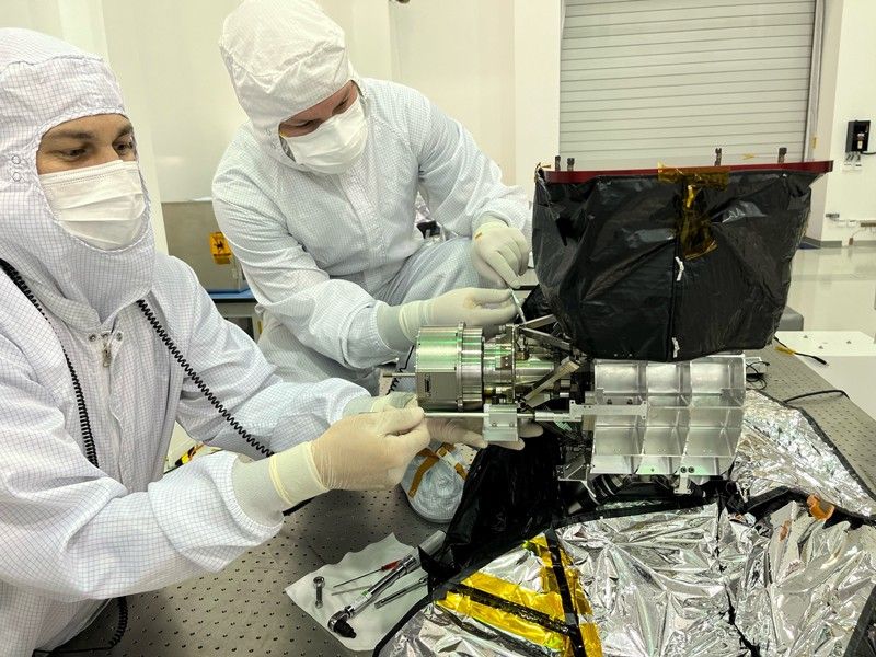 Two engineering flight technicians, one with white and one with light-brown skin, wearing full protective gear at a table in a clean room, installing the flight scanner into the MISE instrument.