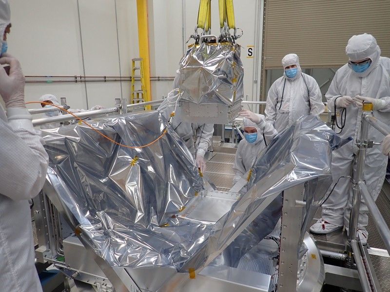 In a sterile white room, five people wearing white full-body coveralls, gloves, and masks monitor a metal box about the size of a microwave oven as a crane lowers the box in to a larger hollow metal box about six feet wide.
