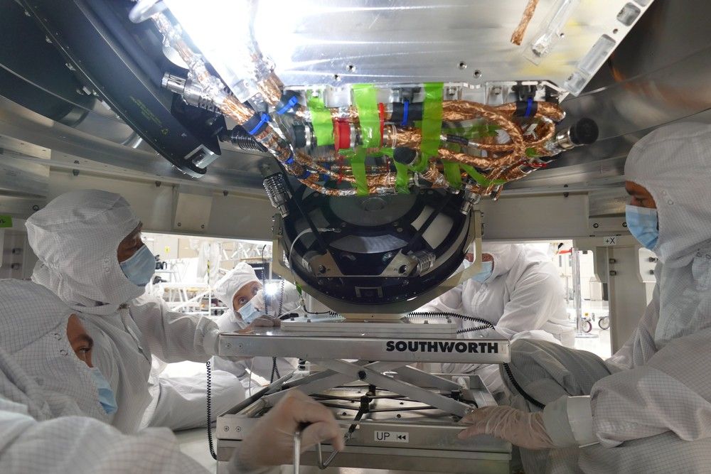 The reaction wheel is seen in the center of this image, shown as a black circulate object two feet wide in width. It’s shown at a slight angle as it is being installed on the spacecraft. Two engineers are seen facing the camera from behind the reaction wheel, and they hold the reaction wheel while it is being installed. The reaction wheel is resting slightly on a small cart with wheels. Above the reaction wheel and the engineers is the body of the spacecraft, which is barely visible.