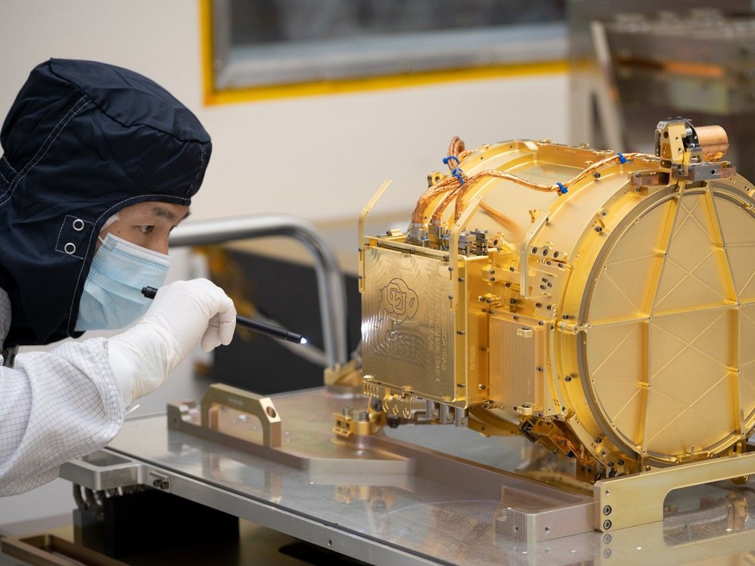 An engineer in a full body coverall and a fask mask is shown on the left side of the image, holding a small black flashlight the size of a pen and examining Europa Clipper’s surface dust analyzer instrument. The instrument is gold colored and about the size of a drum, resting on its side. The instrument is resting upon a silver workbench in the cleanroom.
